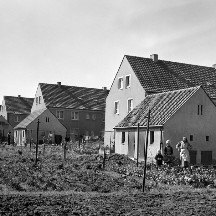 Die schwarz-weiß Fotografie zeigt eine ältere Frau mit zwei Kindern vor einem Haus mit Gemüsegarten. (vergrößerte Bildansicht wird geöffnet)