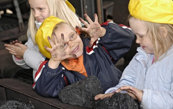 Ein Mädchen mit Helm am Leseband zeigt einem anderen Kind seine mit Kohle verschmutzten Hände.