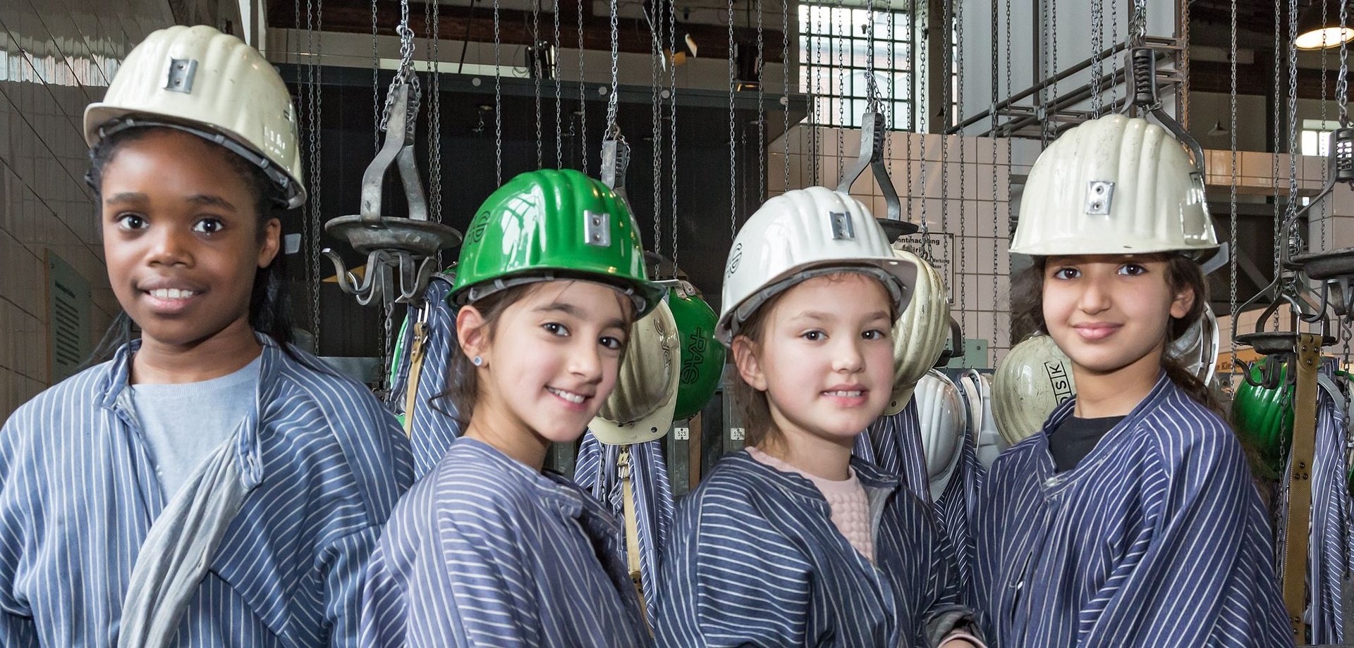 4 Mädchen in Bergarbeiterkleidung und Helmen lachen in die Kamera.