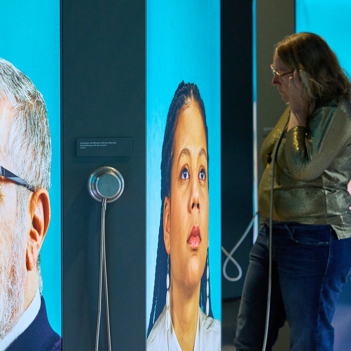 Frau steht vor digitalen Wandpaneelen mit Portraits verschiedener Personen (vergrößerte Bildansicht wird geöffnet)