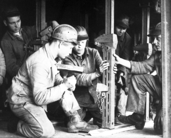 Historisches Schwarz-Weiß-Foto aus den 1950er Jahren mit Lehrlingen und einem Ausbilder bei der Arbeit mit einem Hammer.
