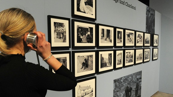 Eine Frau an einer Hörstation vor einer Ausstellungswand mit Fotografien. Eine Frau an einer Hörstation vor einer Ausstellungswand mit Fotografien.