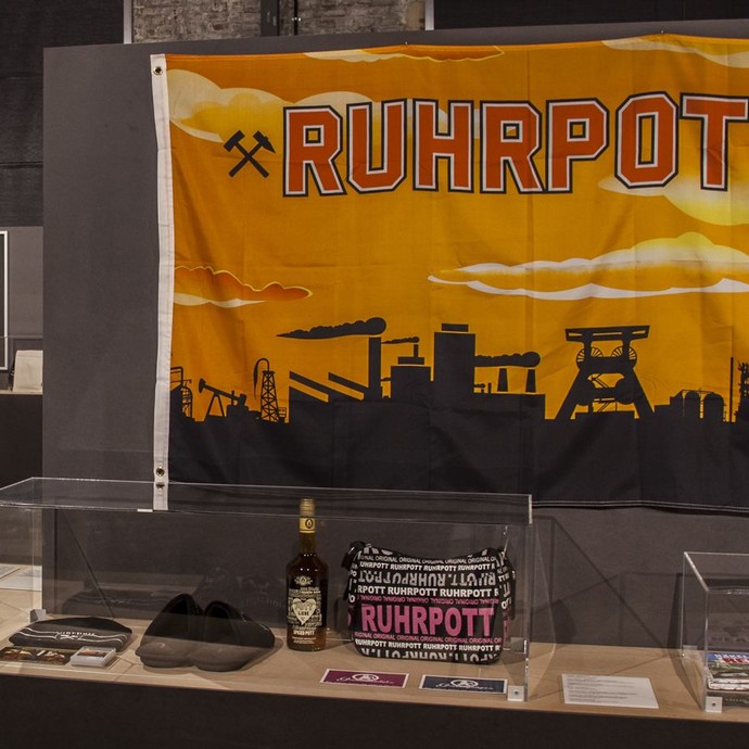 Vitrine mit Ruhrgebietsouvenirs; an der Wand eine große Fahne mit der Aufschrift "Ruhrpott" und typischer Ruhrgebiets-Skyline. (vergrößerte Bildansicht wird geöffnet)