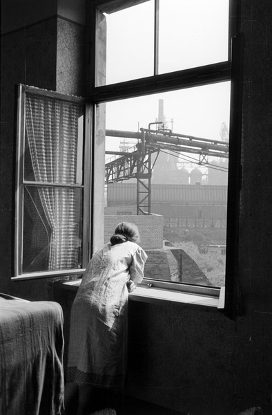 Eine Frau blickt aus dem Fenster ihrer Wohnung in Duisburg-Laar auf die Industrieanlagen der August-Thyssen-Hütte, 1952. Foto: RVR