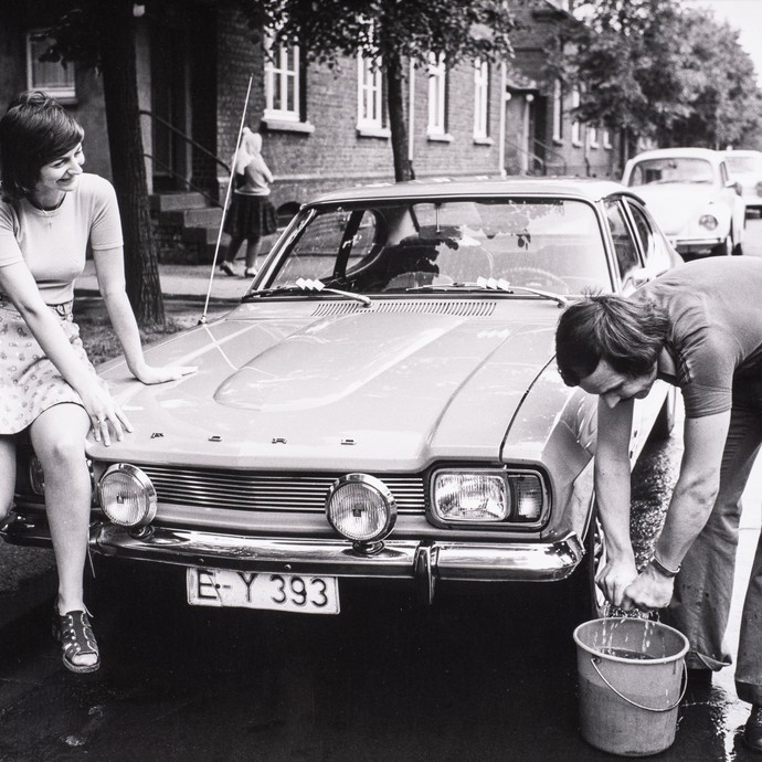 Die schwarz-weiß Fotografie zeigt einen Mann, der ein Auto wäscht und eine Frau, die ihm dabei zu sieht. (öffnet vergrößerte Bildansicht)